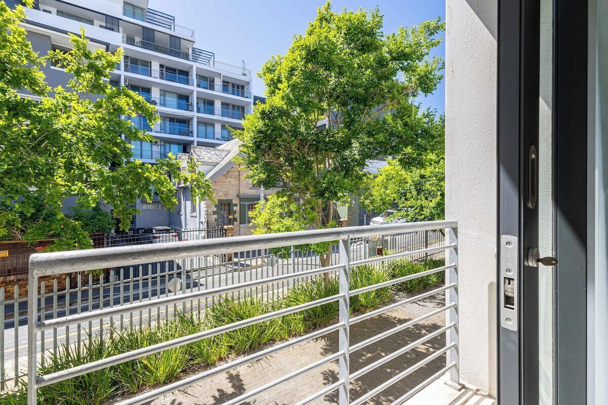 Een balkon met een metalen hekwerk, uitzicht op een meerhoog gebouw en enkele groenvoorzieningen.
