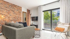 A modern living room with a brick wall, a sofa, a TV, a small round table, and a balcony with a wicker chair. - One77 on Main (Green Point)