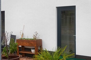 Une jardinière en bois, ornée de plantes, est placée contre un mur blanc, à côté d’une porte vitrée.