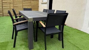 Mesa rectangular al aire libre con sillas, rodeada por una valla de madera, sobre césped artificial.