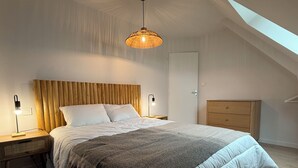Un dormitorio con techo inclinado, una cama con cabecera de madera, dos lámparas de noche, una mesita de noche, un aparador y una lámpara colgante.