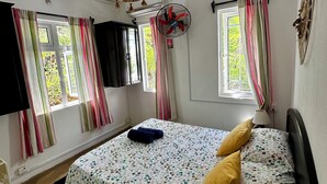 Une chambre à coucher avec un lit, deux fenêtres avec des rideaux, un ventilateur sur pied et un objet décoratif fixé au mur.