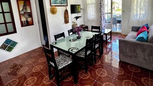 Un coin repas comprenant une table à plateau en verre, des chaises noires, un lustre, un tableau et un canapé avec des coussins.