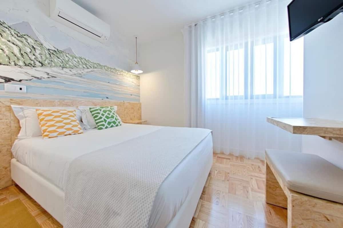 A bedroom with a bed, a TV, a window with curtains, and a wall-mounted air conditioner.