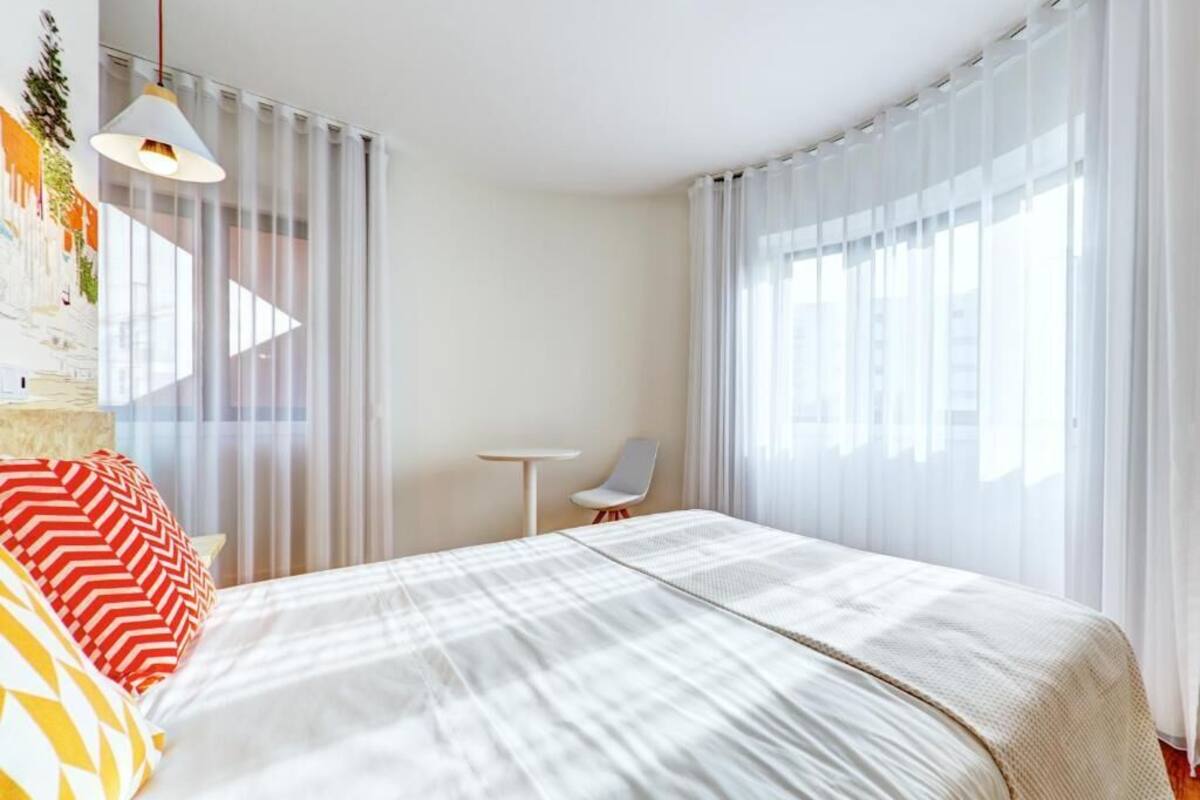 A modern hotel room with a large bed, a small table, a chair, and sheer curtains.