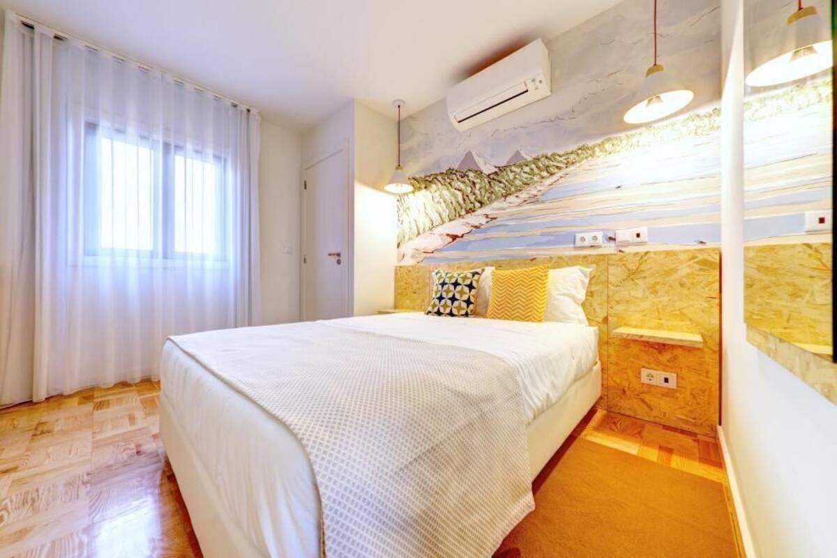 A bedroom with a bed, a window with curtains, an air conditioning unit, and a wall with a mountain mural.