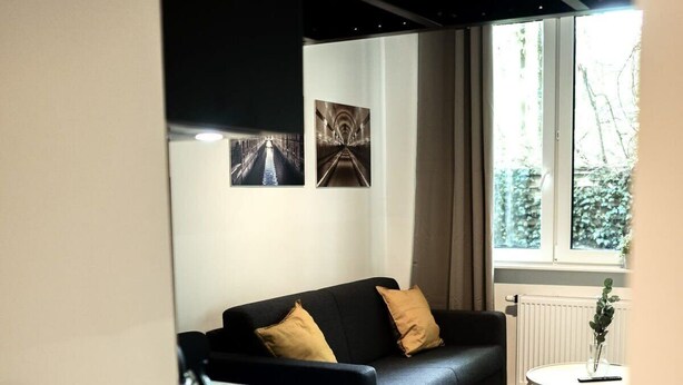 Un salon moderne avec un canapé noir, une petite table ronde et une fenêtre donnant sur des arbres.