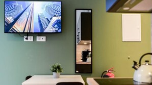 Een kamer met een aan de muur bevestigde televisie, een klein tafeltje met een stoel, een potplant, een brandblusser en een waterkoker op een aanrecht.