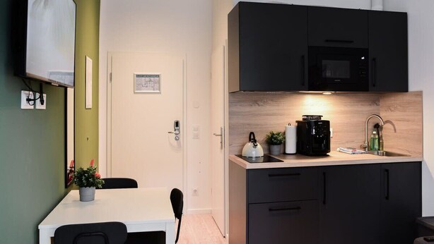 Een kleine keuken met een magnetron, waterkoker en koffiezetapparaat op het aanrecht, een eettafel met stoelen, en een deur met een klein fotolijstje.