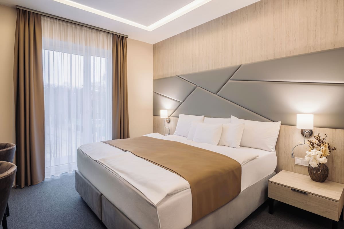 A modern hotel room with a large bed, a nightstand with a lamp, a chair, and a view of the outdoors through sheer curtains.