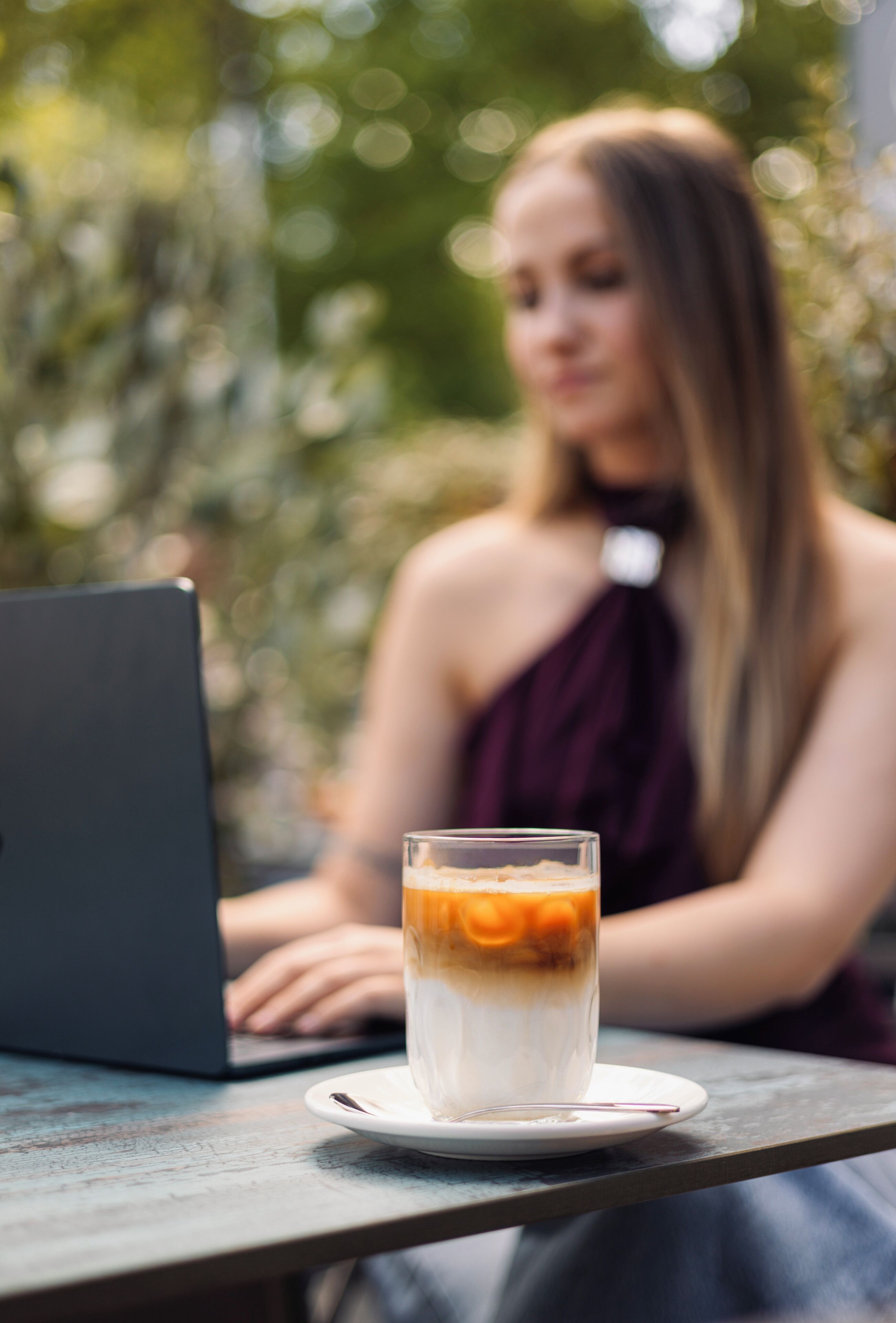 A person using a laptop at an outdoor table with a glass of a layered beverage.