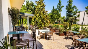 An outdoor patio area with tables and chairs, surrounded by greenery and trees. - Park Hotel (Mosonmagyaróvár)