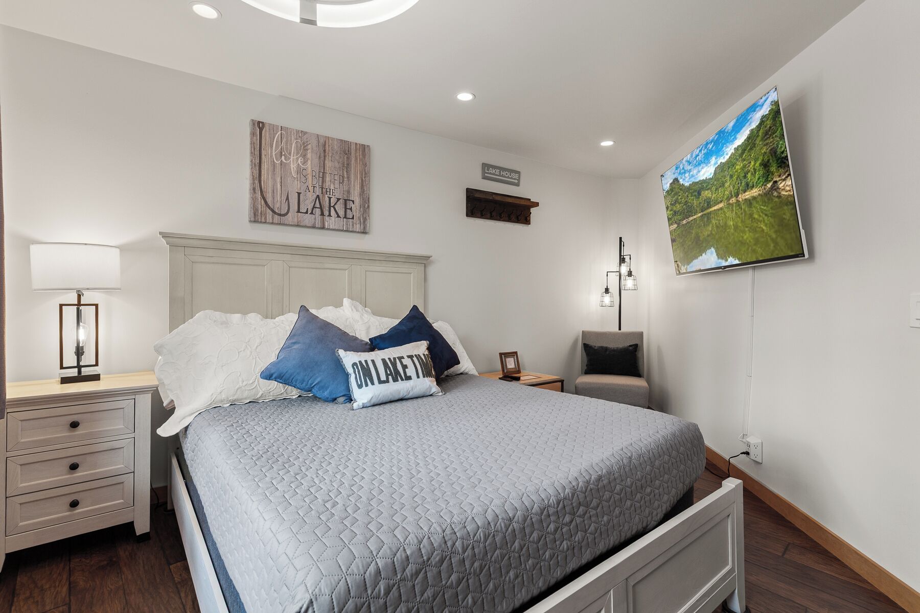 Quarto com cama, mesa de cabeceira, luminária, televisão fixada na parede e placa sobre a cama.