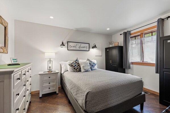 Un dormitorio con cama, mesitas de noche, cómoda y una cita enmarcada en la pared, además de una ventana con cortinas.
