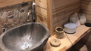 A stone sink with a faucet, a mirror, a stone wall, and wooden shelves with baskets and a white container. - Apartment “Les Gentianes” Valley & Mountain View, 6 Guests, 3 Bedrooms (Chamrousse)