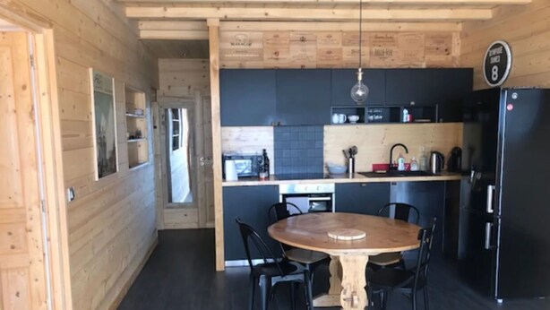 A modern kitchen with dark flooring, wooden walls, a round wooden table with chairs, and a black refrigerator. - Apartment “Les Gentianes” Valley & Mountain View, 6 Guests, 3 Bedrooms (Chamrousse)