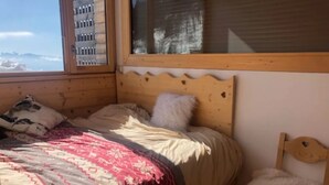 A wooden bed with a patterned blanket, a fluffy pillow, and a view of mountains through a window. - Apartment “Les Gentianes” Valley & Mountain View, 6 Guests, 3 Bedrooms (Chamrousse)
