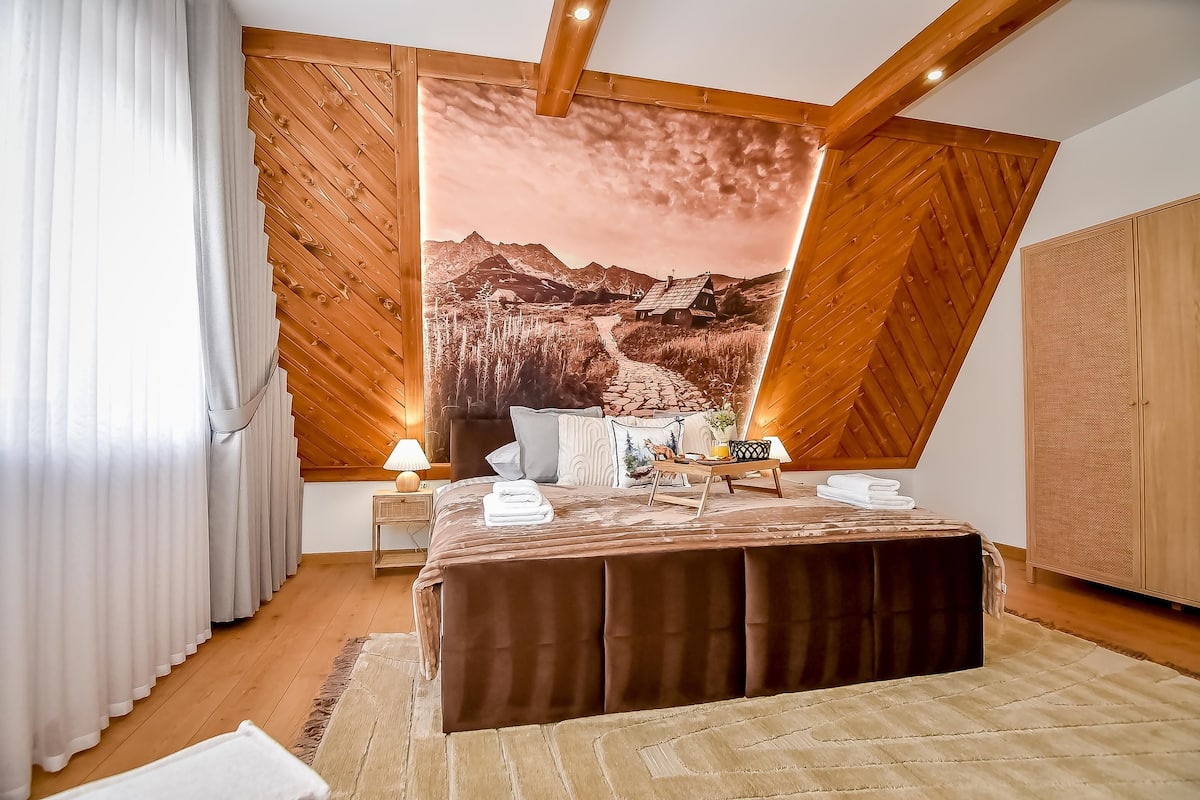 A bedroom with a large bed, wooden ceiling beams, a nightstand with a lamp, and a large wall mural of a mountain landscape.
