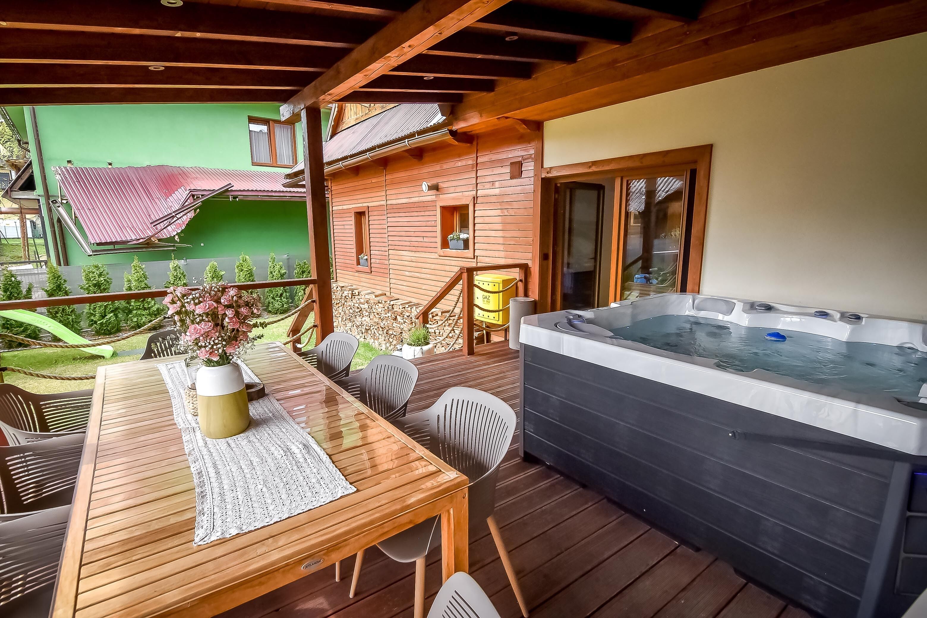 Une terrasse en bois avec un jacuzzi, une table et des chaises, et un petit bâtiment avec un toit rouge.