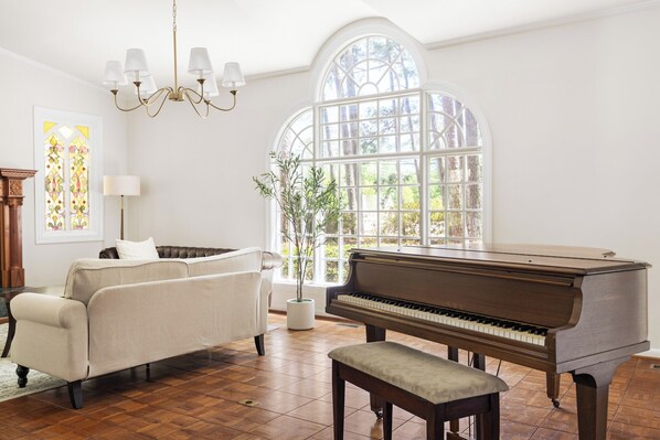 A room with a piano, a sofa, a chandelier, and a large window with a view of trees.