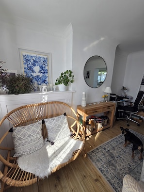 A living room with a wicker chair, a white fireplace, a blue floral painting, and a round mirror. - Room-cozy Guest Room in Vibrant Brighton (Brighton)