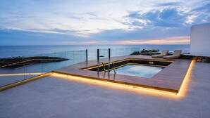 Una piscina en la azotea con vistas al mar, barandilla de cristal y sillones de descanso.