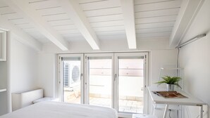 Un dormitorio con cama, escritorio, silla, planta en maceta y puerta corrediza de vidrio que da a un área exterior.