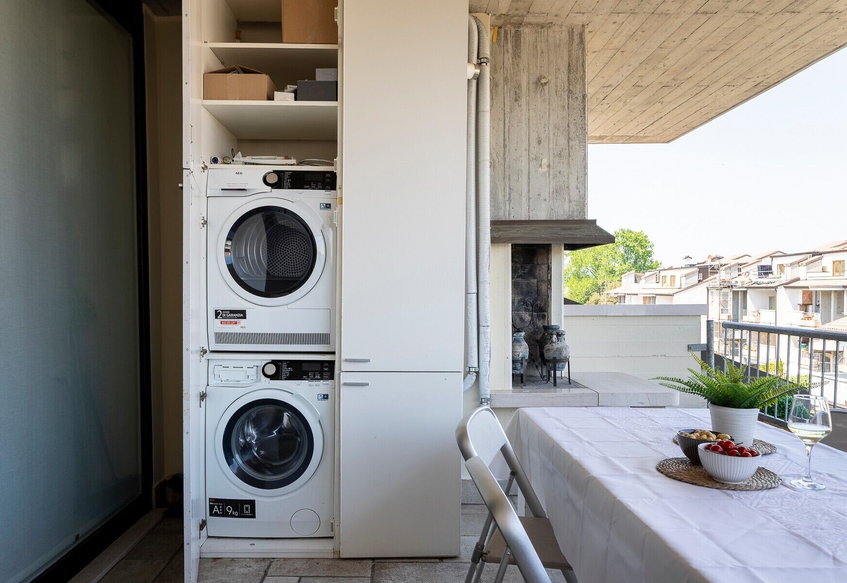 A laundry room with stacked washing machines, a table with a plant and bowls, and a view of buildings in the background.