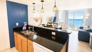 Een moderne keuken met een zwart aanrecht, houten kastjes en een vaatwasser. Er is een zithoek met een tafel en stoelen, en uitzicht op de zee door grote ramen.