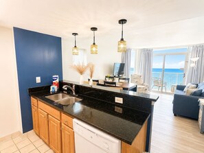 A modern kitchen with a black countertop, wooden cabinets, and a dishwasher. There is a dining area with a table and chairs, and a view of the sea through large windows. - BEACH DAZE GETAWAY/PRIME LOCATION (Myrtle Beach)