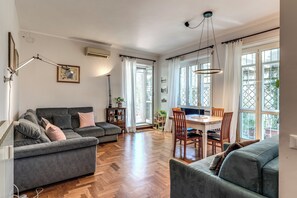 Ein geräumiger Wohnbereich mit grauem Sofa, einem Esstisch mit Stühlen und großen, mit Vorhängen versehenen Fenstern.