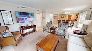 Ein Wohnzimmer mit Fernseher, zwei Sesseln, einer Couch, einem Couchtisch und einem Essbereich im Hintergrund.