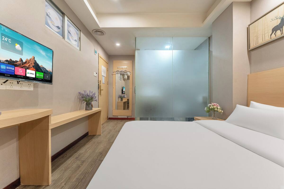 A hotel room with a bed, a TV mounted on the wall, a wooden desk, and a mirror.