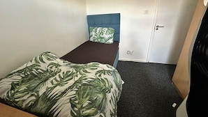 Ein kleines Zimmer mit einem Einzelbett, blauem Kopfteil, dunklem Teppich und weißer Tür.