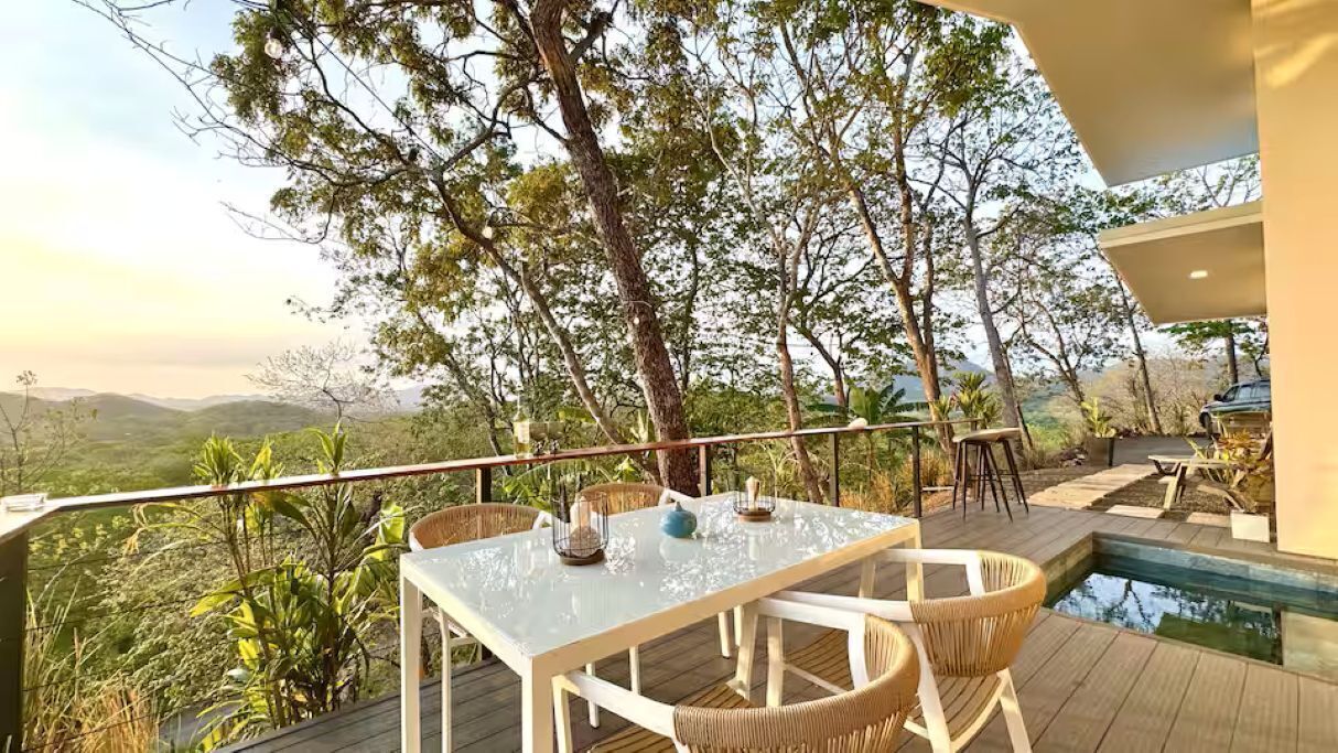 Un espace extérieur aménagé pour les repas, avec une table et des chaises, entouré d’une piscine et offrant une vue sur un paysage montagneux.