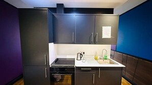 Una cocina compacta con muebles oscuros, horno empotrado, lavamanos y hervidor.