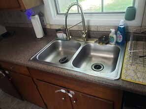 A kitchen with a double stainless steel sink, a window above, and a countertop with a dish rack and cleaning supplies. - Pete's Place  (Tulsa)