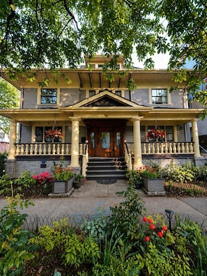 A two-story house with a front porch, steps, and a garden with flowers and plants.