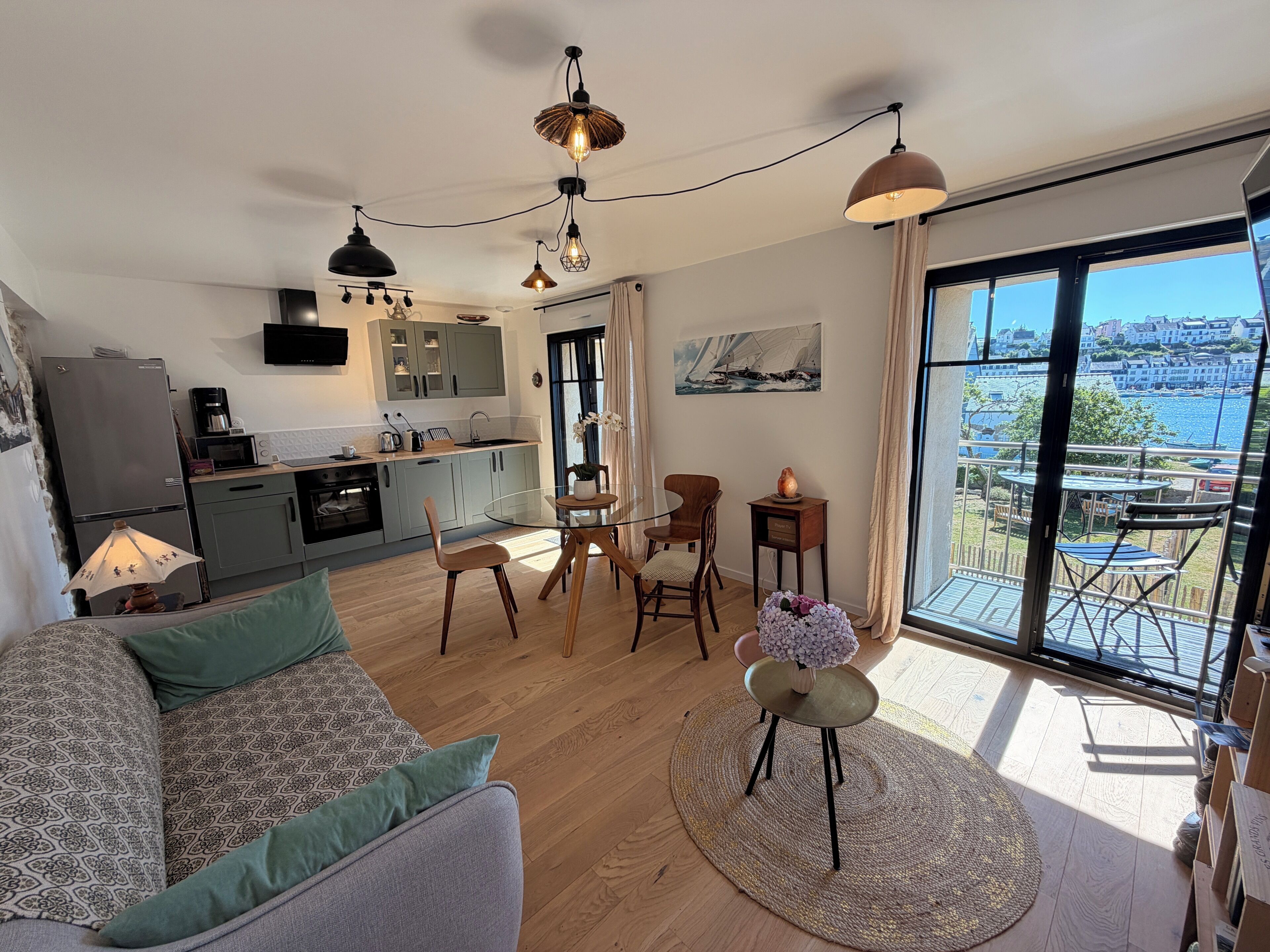 Ein modernes Wohnzimmer mit einer Couch, einem Esstisch mit Stühlen, einer Küchenzeile mit Schränken und Geräten sowie einem großen Fenster mit Meerblick.