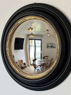 A round mirror with a gold frame reflecting a dining area with a table, chairs, and a hanging light fixture. - Port & sea view - Elegant apartment for 2 in Audierne - L'Oasis sur mer (PLOUHINEC)