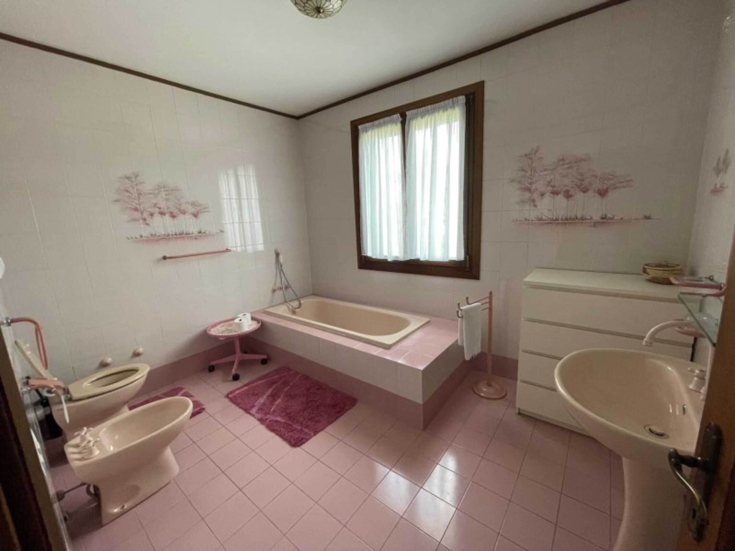 A bathroom with a bathtub, two toilets, a sink, and a window with curtains.
