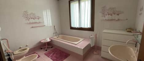 A bathroom with a bathtub, two toilets, a sink, and a window with curtains.