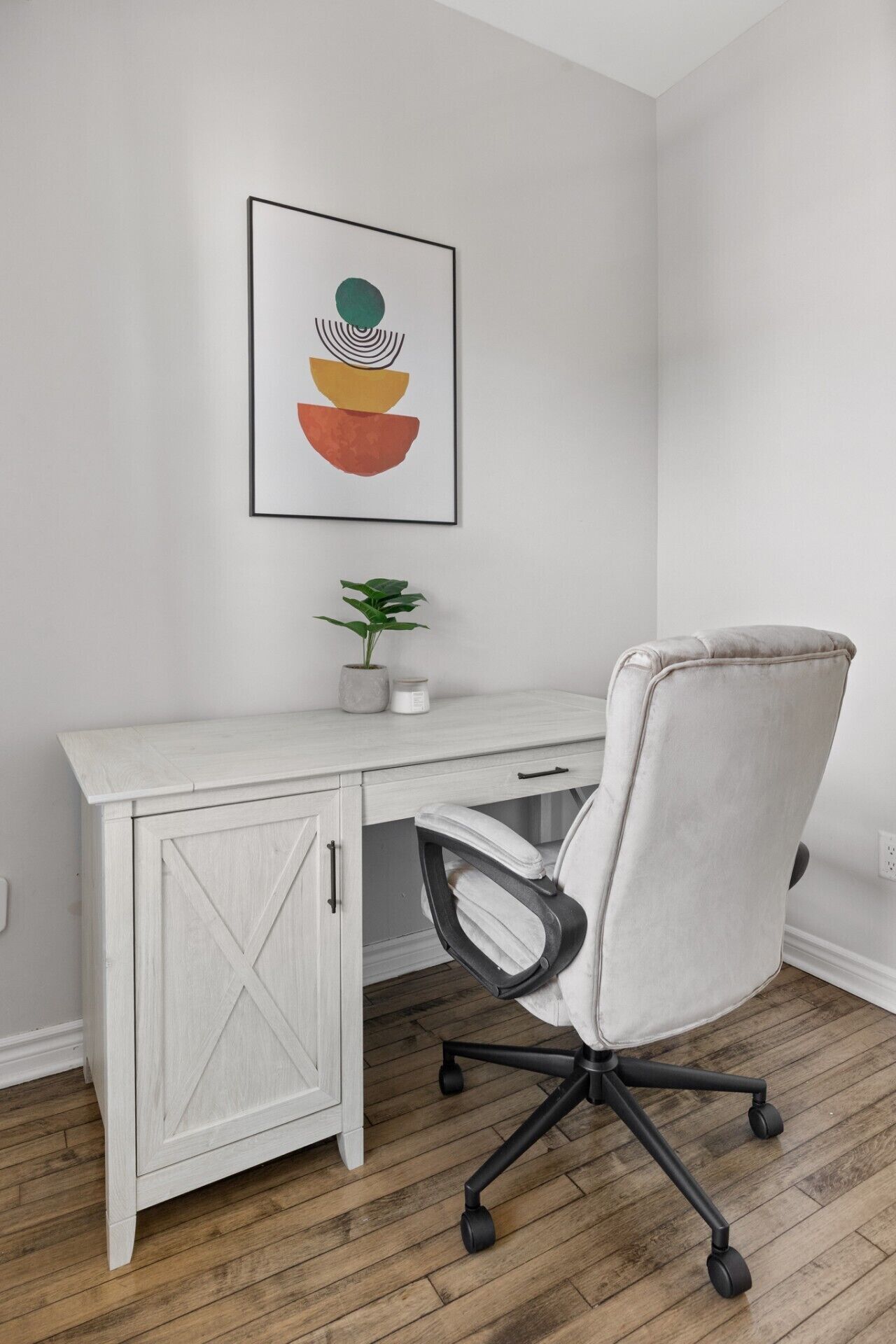 A white desk with a drawer, a cushioned office chair, a potted plant, and a framed abstract artwork on the wall.