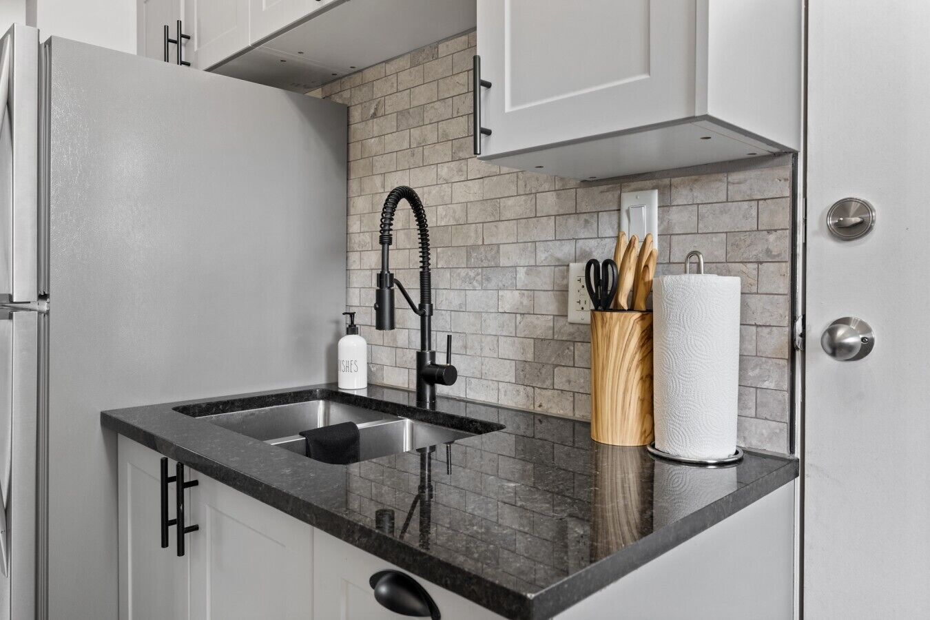 Una cocina con encimera negra, pileta de acero inoxidable y un portalapiedras de madera.