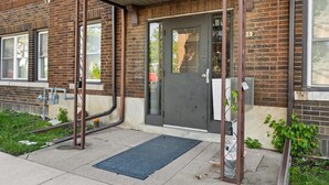 A brick building with a dark door, a metal gate, and a blue mat at the entrance. - Stay Together but with  Privacy of Two Fully Equipped Apartments (Windsor)