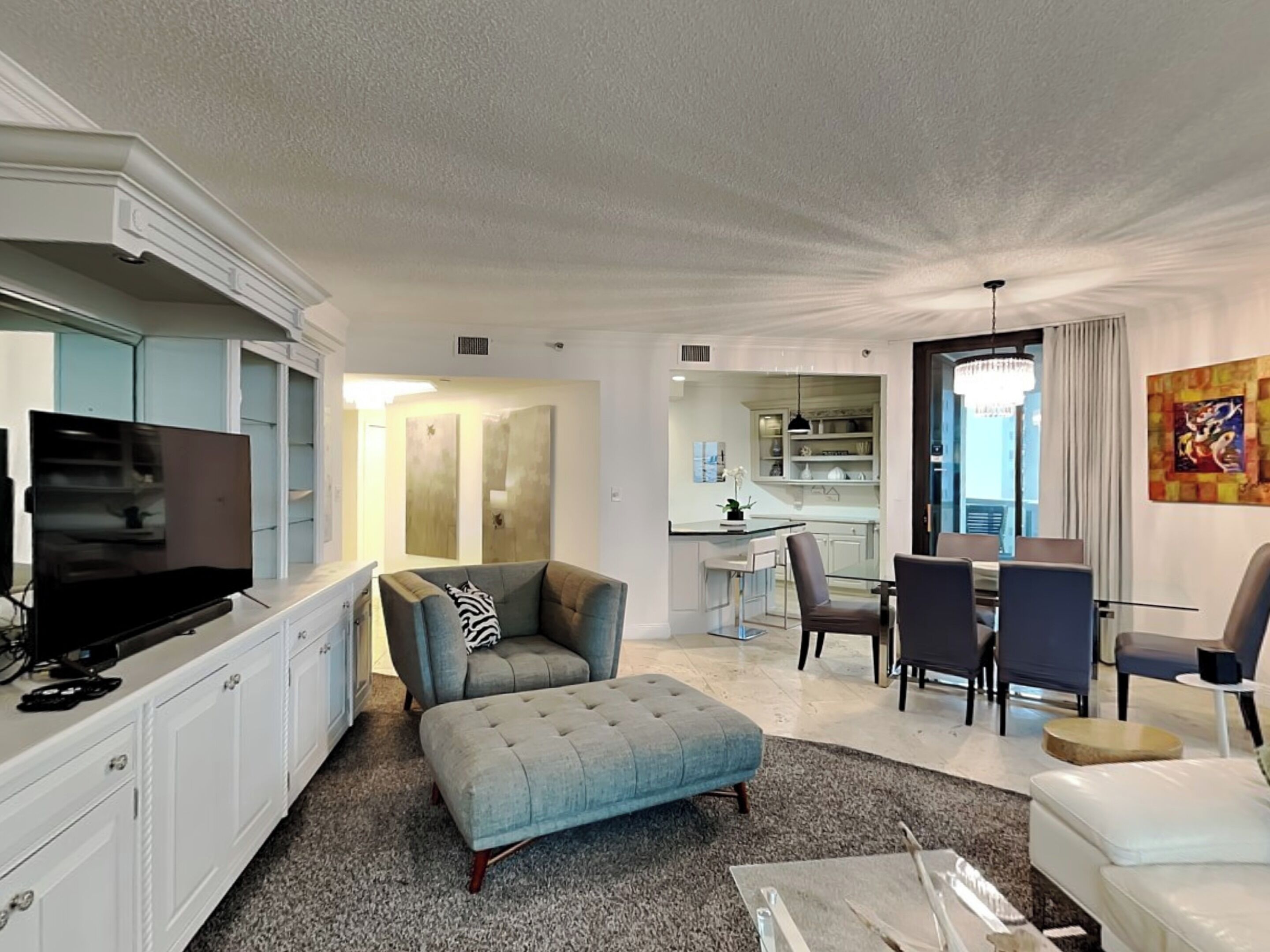 A modern living room with a flat-screen TV, a grey sofa, a dining area with chairs, and a kitchen in the background.