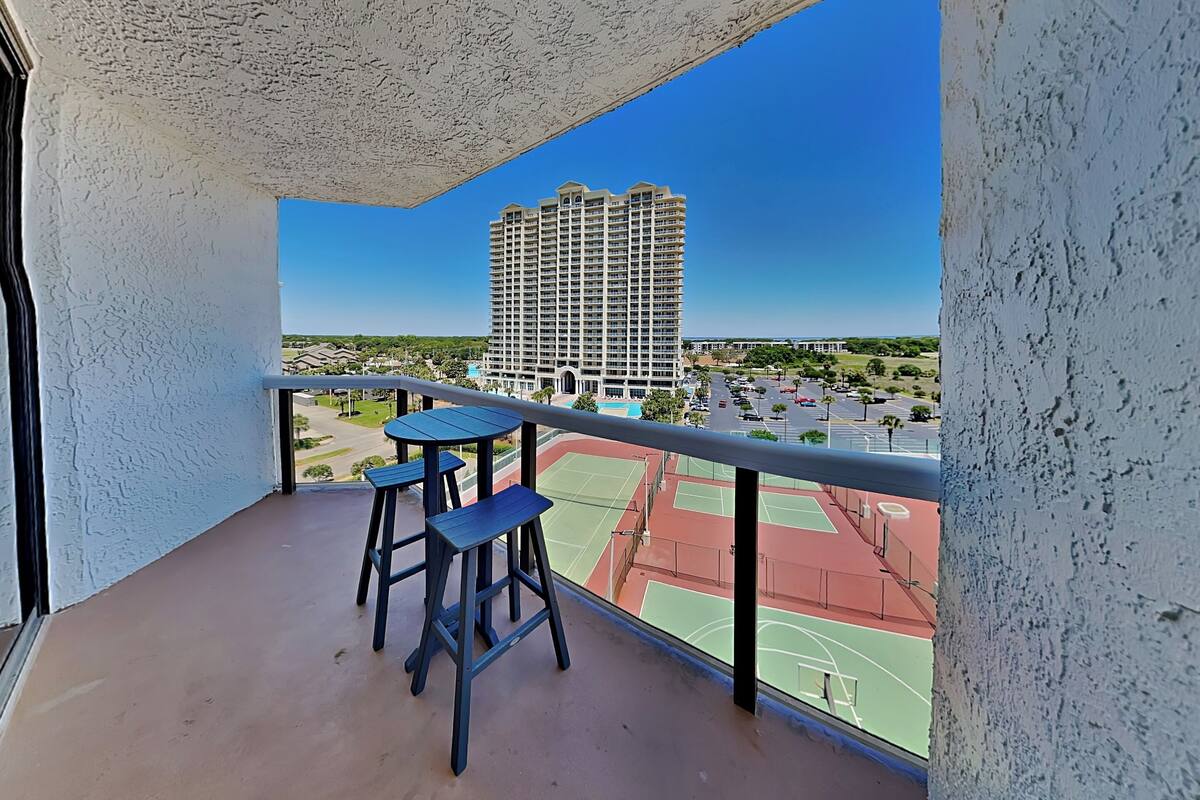 A balcony with a view of a high-rise building, a tennis court, and a parking area.