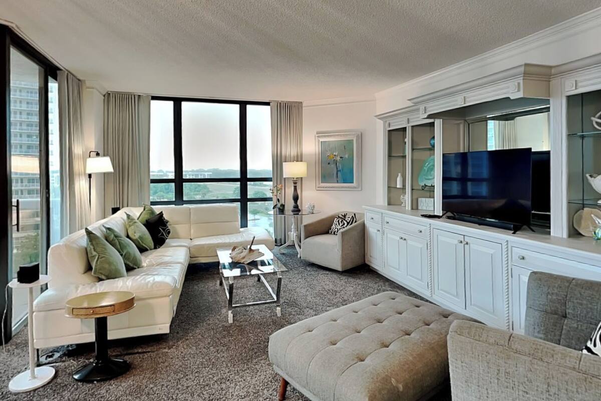 A hotel living room with a sofa, armchair, ottoman, coffee table, TV, and a large window.