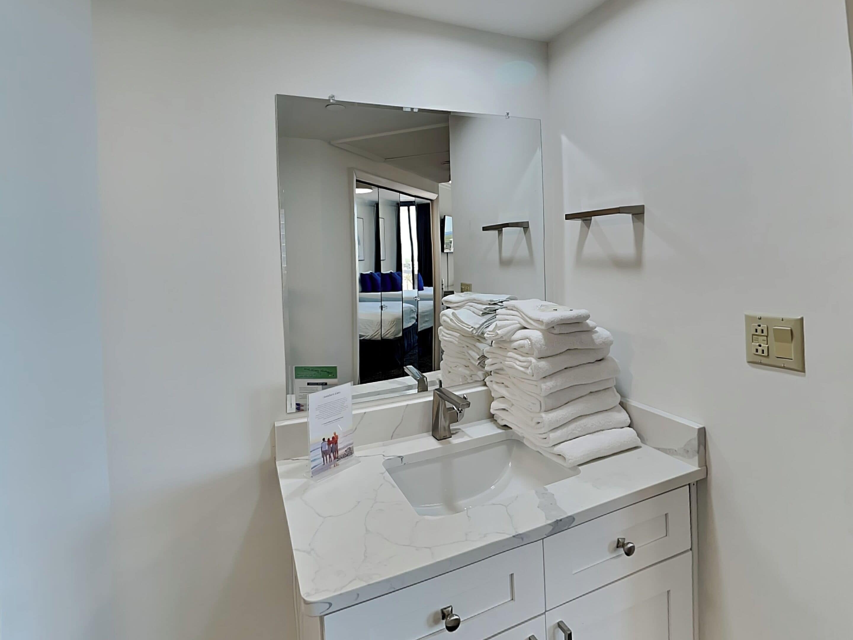 A bathroom with a white marble sink, a mirror, a stack of white towels, and a visible door leading to a bedroom.