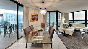 A modern hotel room with a glass dining table, a sofa, a chandelier, and a large window offering a city view. - Surfside #612 & 612a (Miramar Beach)